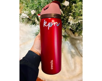 May include: A metallic red Owala water bottle featuring a dark brown lid with a pink carry loop. The bottle displays the text 'owala' in white near the base and the custom script lettering 'kpn' in a light blue and white gradient on the upper body.