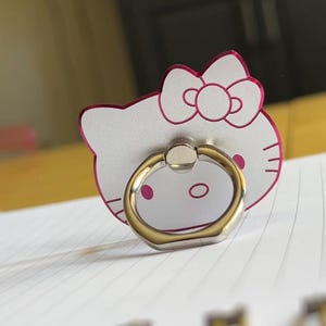 以下が含まれることがあります： ピンクの輪郭を持つハローキティのデザインのシルバーの電話リング。リングは金色のバンドで、キャラクターの頭の後ろに取り付けられています。リングは、電話グリップまたはスタンドとして使用するように設計されています。