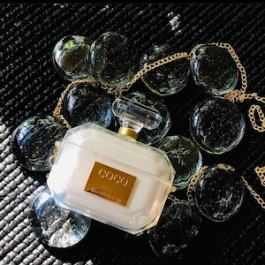 May include: A clear plastic phone case shaped like a bottle of perfume. The case has a gold chain attached to it and features the text "COCO" and "PARIS" on the front. The case is sitting on a black surface with clear glass beads.
