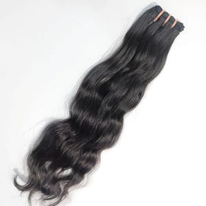 Puede incluir: Una extensión de cabello larga y ondulada de color negro intenso. El cabello está sujeto a una banda de tela reforzada en la parte superior, con una textura consistente y un brillo natural que cae en ondas suaves a lo largo de toda la pieza.