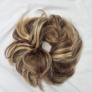 Op de afbeelding: Een cirkelvormige haarextensie in de vorm van een scrunchie met een mix van lichtblonde en middenbruine highlights. Het synthetische haar is gestyled in een rommelige, volumineuze knot met losse plukken en een textuur voor volheid.