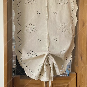 Könnte beinhalten: Cremefarbener, bestickter Fenstervorhang mit gewelltem Rand. Der Vorhang hat florale und blattförmige Ausschnitte und ist hochgebunden, um das dahinterliegende Fenster freizugeben. Der Vorhang hängt an einer Holzstange über einer Holztür.