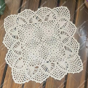 Peut inclure: Nappe en crochet faite à la main, de couleur crème, avec un motif floral et géométrique. La nappe a un bord festonné et est posée sur une surface en bois. La nappe mesure environ 30 cm de diamètre, idéale pour un centre de table ou un élément décoratif.