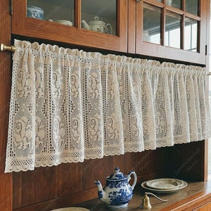 May include: A cream-colored lace cafe curtain featuring a repeating floral pattern and a scalloped bottom edge. The curtain is gathered on a brass rod and hangs in front of a wooden cabinet with glass panels, displaying a blue and white teapot.