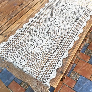 Puede incluir: Camino de mesa de ganchillo de tejido abierto color crema. El camino de mesa presenta un borde festoneado y un patrón floral repetitivo en el centro. Se muestra sobre una mesa de madera rústica.