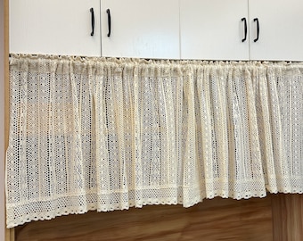 May include: A cream-colored crocheted valance with a delicate pattern hangs over a set of white kitchen cabinets. The valance has a scalloped edge and is made of a lightweight, airy fabric.