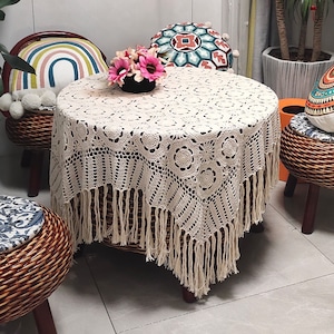 以下が含まれることがあります： フリンジが付いた白いクロシェのテーブルクロスで、繊細な花柄が特徴です。テーブルクロスは、籐製の椅子とカラフルなクッションのある丸いテーブルにかけられています。