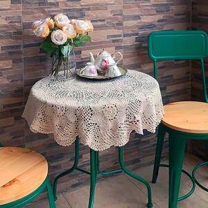Könnte beinhalten: Ein runder Tisch mit einer beigen gehäkelten Spitzentischdecke, darauf eine klare Glasvase mit pfirsichfarbenen künstlichen Rosen, eine weiße Porzellanteekanne und zwei passende Teetassen auf einem Tablett, flankiert von zwei grünen Metallstühlen.