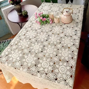 Op de afbeelding: Een wit gehaakt tafelkleed met een delicaat bloemenpatroon. Het tafelkleed ligt op een houten tafel met een theepot en kopjes erop.