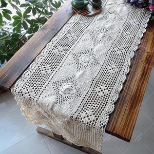 May include: A white crocheted table runner with a delicate floral pattern. The runner is laid on a wooden table and has a scalloped edge.