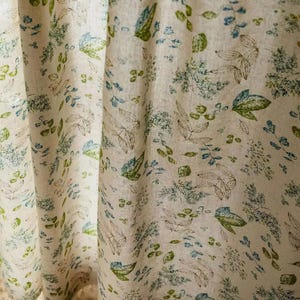 May include: A beige fabric with a repeating pattern of green and blue leaves. The fabric has a subtle texture and a light, airy feel.