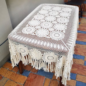Puede incluir: Mantel rectangular de ganchillo color crema con un patrón floral y flecos. El mantel está drapeado sobre una mesa de madera, mostrando el intrincado diseño calado. Los flecos cuelgan a lo largo de los bordes.
