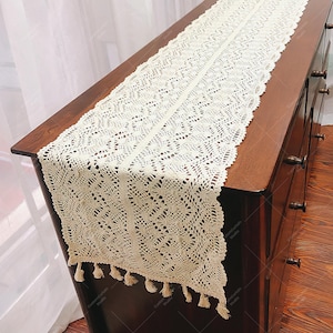 May include: A white crocheted table runner with a delicate floral pattern and fringe trim. The runner is laid on a dark wood surface.