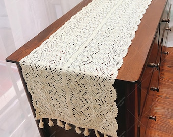 Può includere: Un runner da tavolo in crochet bianco con un delicato motivo floreale e bordi a frangia. Il runner è appoggiato su una superficie in legno scuro.