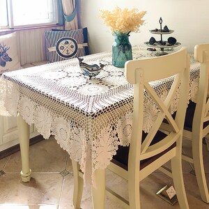 May include: A rectangular dining table covered with a white crocheted lace tablecloth featuring intricate floral patterns and a scalloped edge. The table is accompanied by two cream-colored wooden chairs with an X-shaped back design.