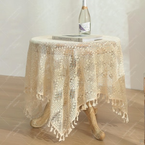 Puede incluir: Una pequeña mesa redonda de madera cubierta con un mantel de ganchillo crema de tejido abierto. El mantel tiene un ribete de flecos. Una botella y un libro titulado "Simplicity at Home" descansan encima.