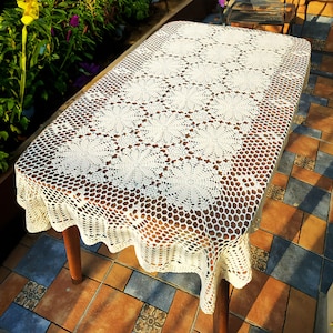 May include: A rectangular off-white crochet tablecloth featuring a repeating pattern of circular floral motifs connected by an openwork hexagonal mesh border that drapes over the edges of a wooden table with four legs.