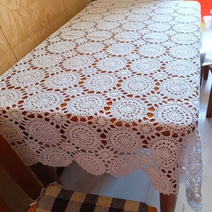 Może przedstawiać: Biały obrus szydełkowy z delikatnym wzorem kwiatowym. Obrus jest położony na drewnianym stole.