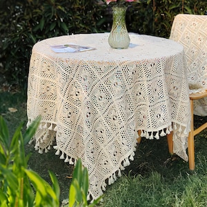 May include: A white crocheted tablecloth with fringe trim, draped over a round wooden table with a vase of pink flowers on top. The table is set in a garden setting with green grass and plants.