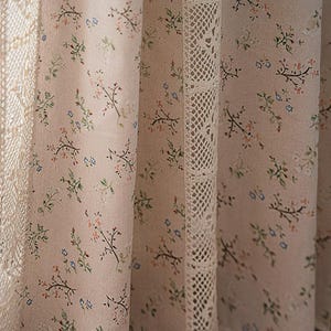 Può includere: Una coppia di tende trasparenti con un motivo floreale e un bordo in pizzo bianco. Le tende sono di colore beige chiaro con piccoli fiori rosa, blu e verdi.