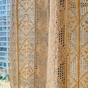 Op de afbeelding: Een close-up van een beige gehaakt gordijn met een delicaat patroon. Het gordijn is gemaakt van een fijn, lichtgewicht materiaal en heeft een zacht, luchtig gevoel.