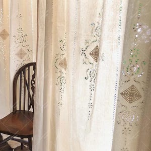 Op de afbeelding: Witte, doorschijnende gordijnen met ingewikkelde geborduurde ontwerpen met kleine spiegelaccenten. De gordijnen hangen in een kamer met een houten stoel op de voorgrond.