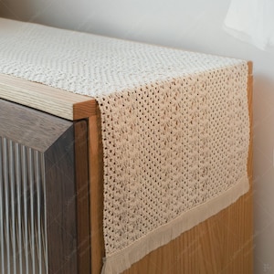 May include: A cream-coloured crochet table runner with a fringed edge, placed on a wooden cabinet. The runner's open weave pattern is evident, and the cabinet has a light wood top and a darker wood body.