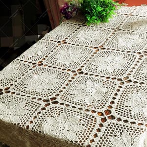Puede incluir: Un mantel de crochet blanco con un patrón cuadrado y acentos florales. El mantel está colocado sobre una mesa de madera.