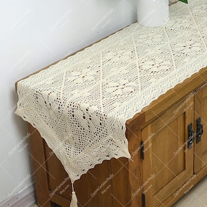 Puede incluir: Un camino de mesa de ganchillo con un delicado patrón floral en color crema. El camino de mesa tiene un borla en un extremo y está colocado sobre una mesa de madera.