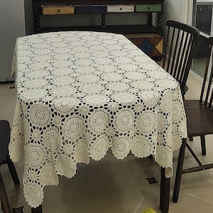 May include: A rectangular off-white crochet tablecloth featuring a repeating pattern of circular motifs connected by smaller star-shaped elements. The fabric has an openwork design with scalloped edges that drape over the sides of a table.