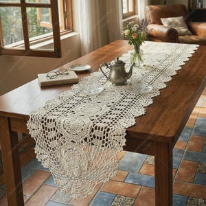 May include: A cream-colored crochet table runner with an intricate floral pattern, featuring a metallic teapot, two matching teacups on saucers, a small book, and a pair of reading glasses arranged on a wooden table surface.