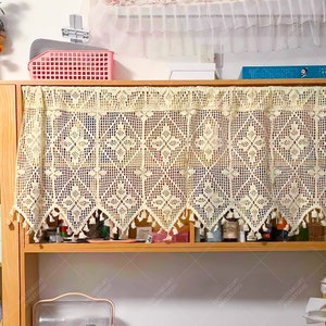 May include: A crocheted valance with a scalloped edge and fringe trim hangs over a wooden shelf. The valance is cream colored and has a delicate floral pattern.