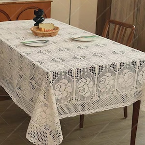 Pode incluir: Toalha de mesa retangular creme com um padrão floral de renda. A toalha de mesa está sobre uma mesa de madeira, com uma cesta de pão e dois pratos visíveis. O desenho de renda apresenta rosas e formas geométricas.