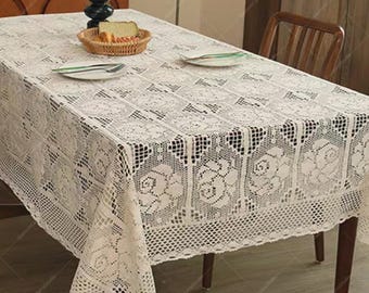 May include: A rectangular off-white lace tablecloth featuring a repeating floral rose pattern and a decorative crocheted border. The fabric is draped over a table, with two white plates, silverware, and a woven basket containing bread.