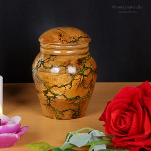 May include: A wooden urn with a green and brown woodgrain pattern. The urn has a lid and is handcrafted. The urn is sitting on a wooden surface with a red rose and a purple candle.