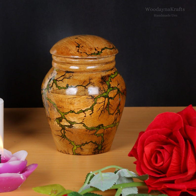 May include: A wooden urn with a green and brown woodgrain pattern. The urn has a lid and is handcrafted. The urn is sitting on a wooden surface with a red rose and a purple candle.