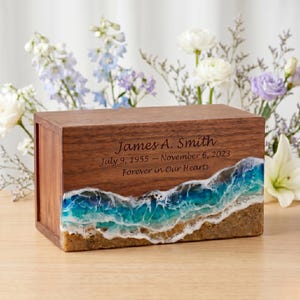 Könnte beinhalten: Eine Holzurne mit einem Strandmotiv. Die Oberseite und die Seiten sind aus dunklem Holz, während der Boden einen sandigen Strand mit blauen und türkisfarbenen Wellen zeigt. Der Text lautet "James A. Smith July 9, 1955 - November 6, 2023 Forever in Our Hearts."