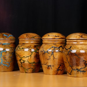 May include: Four wooden jars with lids, each with a unique pattern of blue, green, and brown lines. The jars are arranged in a row on a wooden surface.