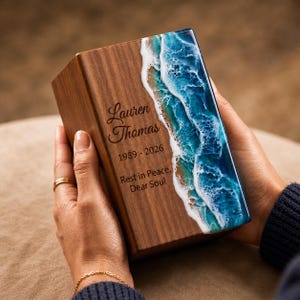 May include: A wooden memorial urn with a resin ocean wave design. The urn is engraved with the name "Lauren Thomas", birth and death years, and the phrase "Rest in Peace, Dear Soul". The urn is held by a person.