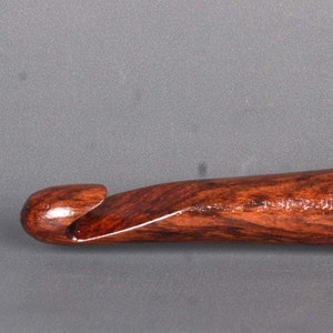 May include: A close-up of a wooden crochet hook with a dark brown and reddish-brown wood grain. The hook is polished and has a smooth finish.