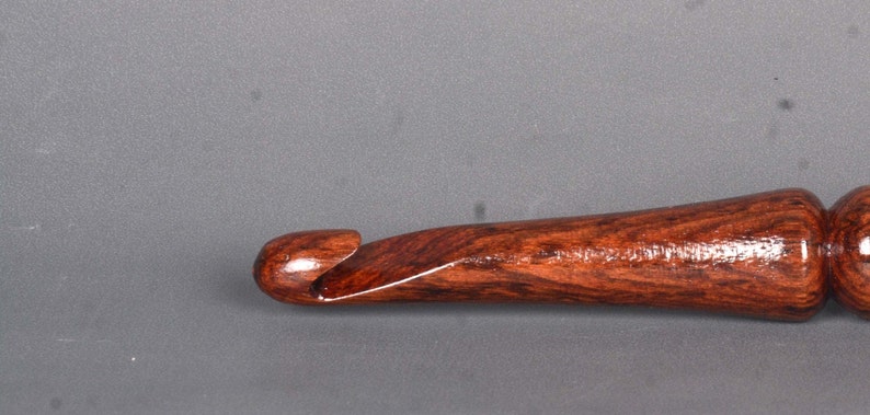 May include: A close-up of a wooden crochet hook with a dark brown and reddish-brown wood grain. The hook is polished and has a smooth finish.