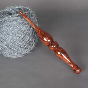 Peut inclure: Une pelote de laine grise à côté d'un crochet en bois artisanal. Le crochet est d'un brun foncé riche avec une finition brillante, avec un manche décoratif. La laine semble douce et duveteuse.