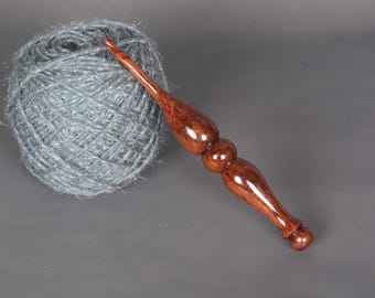 May include: A ball of gray yarn next to a handcrafted wooden crochet hook. The hook is a rich, dark brown with a glossy finish, featuring a decorative handle. The yarn appears soft and fluffy.