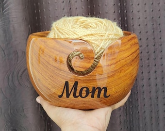 Peut inclure: Un bol à laine en bois avec un motif sculpté et le mot "Maman" gravé sur le devant. Le bol est rempli d'une pelote de laine jaune clair.