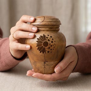 May include: A wooden urn with a sunflower design and the word "love" engraved on the side. The urn is held by two hands. The urn is brown and has a lid.
