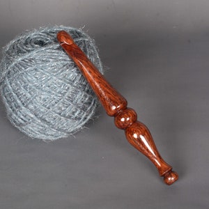 May include: A wooden crochet hook with a dark brown and red wood grain pattern. The hook is lying on a gray surface next to a ball of gray yarn.