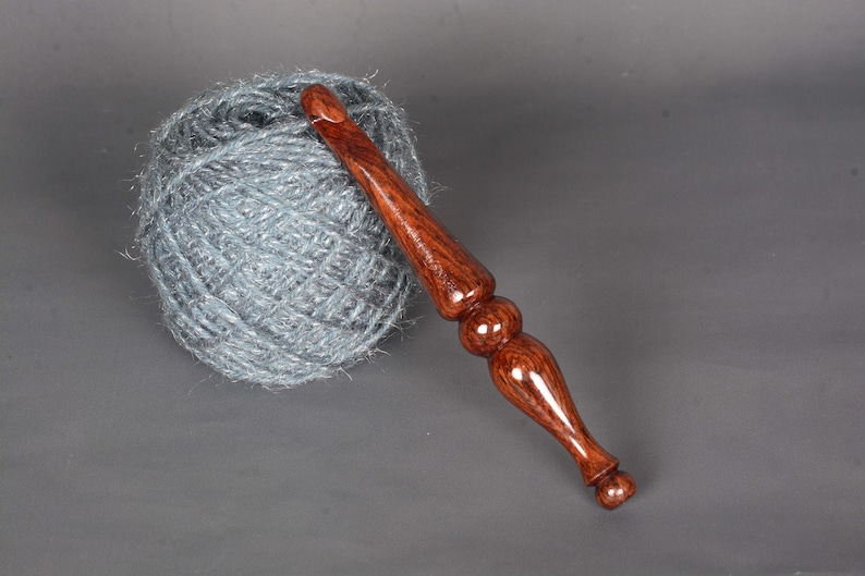 May include: A wooden crochet hook with a dark brown and red wood grain pattern. The hook is lying on a gray surface next to a ball of gray yarn.