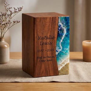 May include: A rectangular wooden cremation urn featuring a dark brown wood grain finish and a vertical side panel of blue and white resin art resembling ocean waves and sandy shore. The front is engraved with Natalie Lewis, 1987-2025, Rest in Peace, Dear Soul.