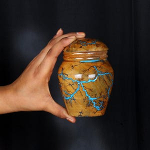 Puede incluir: Un tarro de madera con tapa, decorado con un patrón de remolino azul y marrón. El tarro está hecho a mano y tiene un diseño único y artístico.