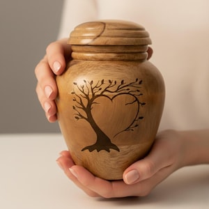 May include: A light brown wooden urn featuring a heart-shaped tree design. The urn has a darker brown tree graphic. The lid is also wooden and rounded. The urn is held in two hands.